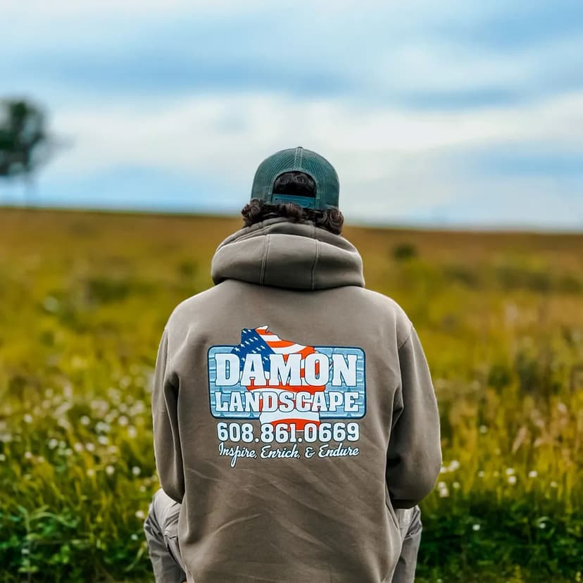 Person wearing a Damon Landscape hoodie in a grassy field, showcasing the company logo and contact.