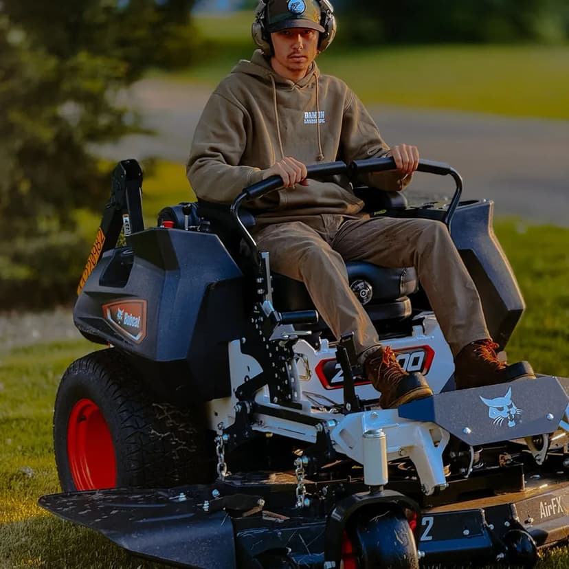 Man operating a zero-turn lawn mower on a grassy area, wearing headphones and casual clothing.